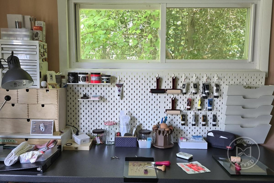 A home printmaking studio with hand-carving tools, inks, and brayers arranged on a white pegboard beside a window of green trees.