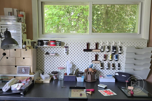 A home printmaking studio with hand-carving tools, inks, and brayers arranged on a white pegboard beside a window of green trees.