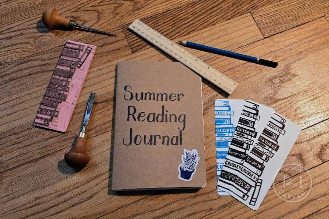 Overhead flatlay of hand-carving tools, a brayer, a pink book-stack stamp block, a kraft Summer Reading Journal, a ruler and pencil, and three book-spine bookmark prints in blue, black, and brown