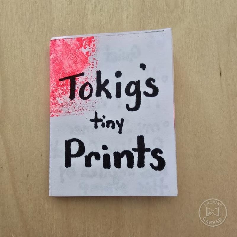 Handmade zine titled 'Tokig's Tiny Prints' resting on a wood surface, showcasing hand-carved eraser stamp prints.